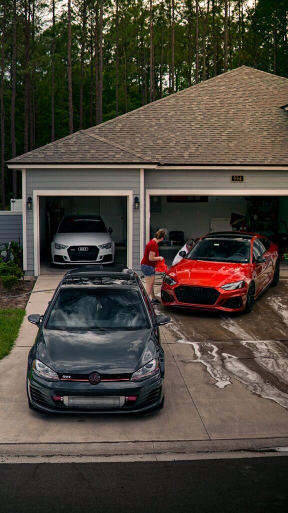 A person washing modern cars in a driveway surrounded by a lush backyard.